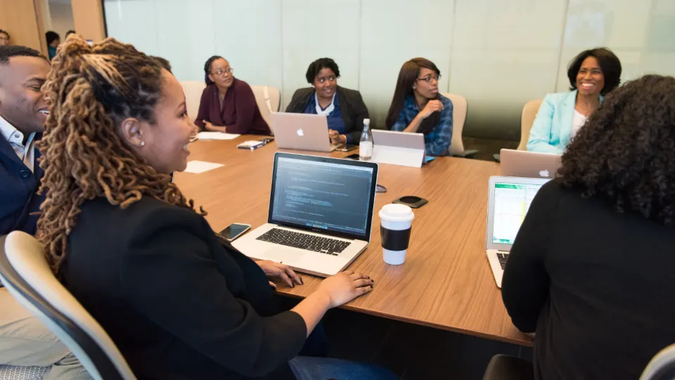 A group of smiling colleagues at a conference table with open laptops